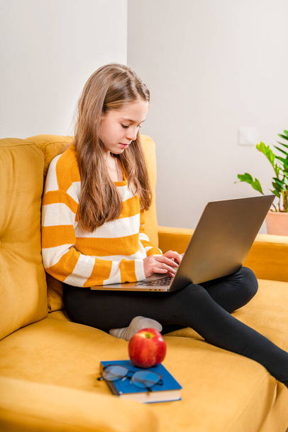 Ein lächelndes europäisches Teenager-Mädchen benutzt einen Laptop für Videoanrufe zu Freunden und Eltern, die auf einem gelben Sofa sitzen. Online-Fernunterricht. - Foto, Bild