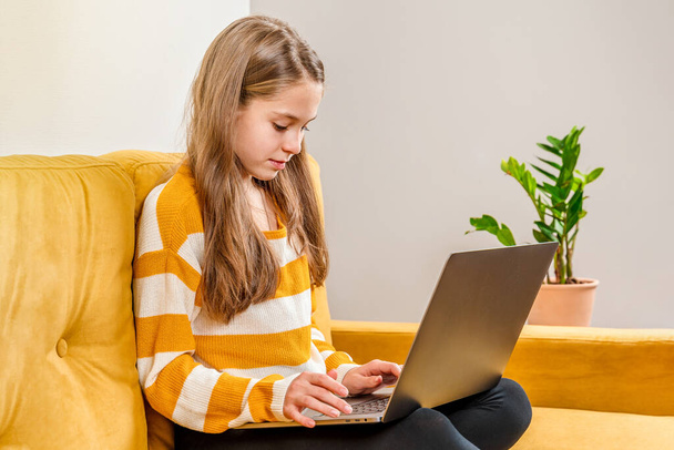 Gülümseyen Avrupalı genç kız, sarı bir kanepede oturan aile ve arkadaşlarına bilgisayar araması yapmak için dizüstü bilgisayar kullanıyor. Uzaklık öğrenimi çevrimiçi eğitim. - Fotoğraf, Görsel