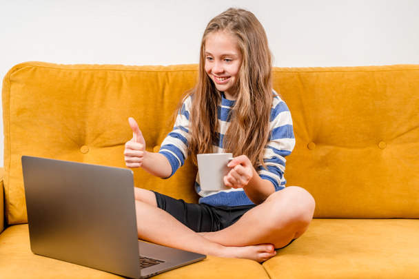 Glimlachend Europees tienermeisje gebruikt een laptop voor videogesprekken naar vrienden en ouders die op een gele bank zitten. Online onderwijs op afstand. - Foto, afbeelding