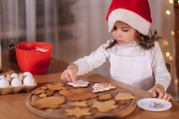 Домашняя пекарня, приготовление традиционных праздничных сладостей. Маленькая девочка в красной шляпе Санты готовится к приготовлению пряничного теста на деревянном столе. Новогодние праздники. Рождественское настроение - Фото, изображение