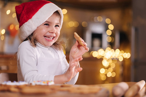 Adorable little girl in santa hat eating homemade gingerbread and laughing. Happy little kid preparing for Christmas. Background of fairy lights bokeh - Фото, изображение