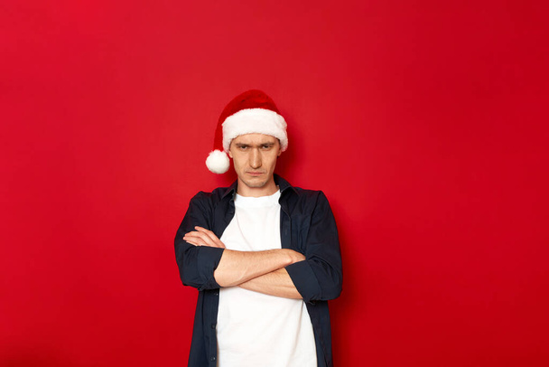 enojado hombre descontento dobló los brazos con la cruz en el pecho y ofendido en Santa Claus sombrero de Navidad. aislado en el espacio de fondo rojo para texto. concepto - personas, emociones negativas, escéptico, odiador - Foto, Imagen