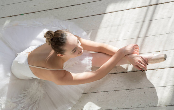 Балерина в белой одежде и юбке лежит с протянутыми руками и длинными ногами на белом деревянном полу - Фото, изображение
