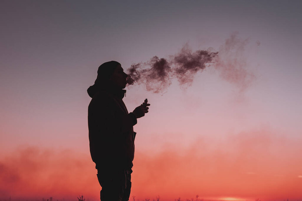 Un hombre con chaqueta fuma un cigarrillo electrónico al aire libre. Vapeando sobre un fondo al atardecer. Alternativa a los cigarrillos analógicos - Foto, imagen