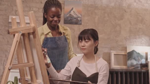 Mediano de joven hombre caucásico y mujer asiática usando delantales, sentado en clase, pintando sobre lienzo en caballete, mentora afroamericana caminando y ayudándoles - Metraje, vídeo