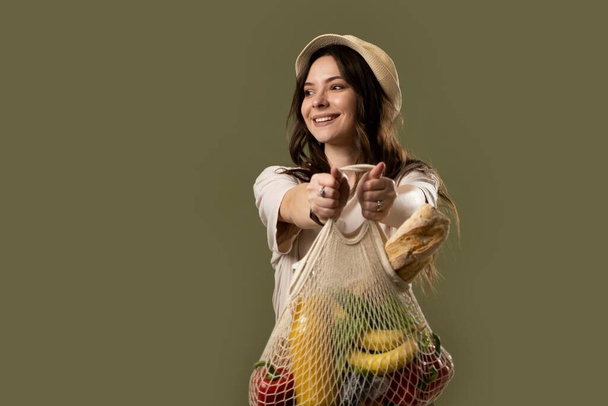 Eco bag string bag with fruits, vegetables, juice and bread in a hands of a brunette girl in a beige suit and a hat. Eco friendly concept. Fashion photography. Zero waste concept. - Foto, imagen
