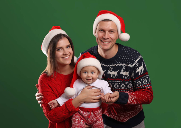 Happy couple with cute baby in Christmas outfits and Santa hats on green background - Fotó, kép