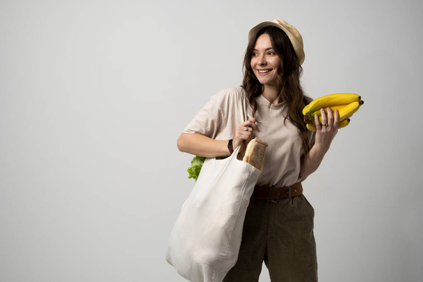 Zero waste concept. Young brunette woman holding cotton eco bag with organic fruits and vegetables on a shoulder and holding bananas in a hand. Using reusable crochet net bag for grocery shopping. - Foto, imagen