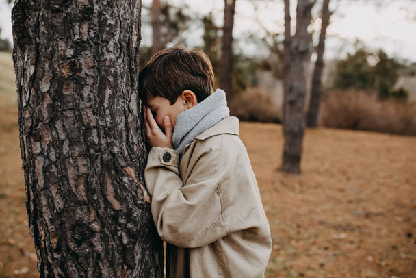 Child playing hide and seek, counting with closed eyes next to a tree in a park. - Фото, изображение