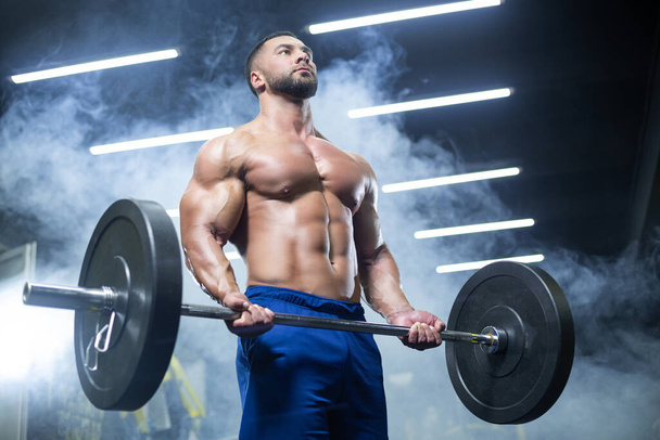 Bottom up view of a muscular athlete lifting heavy barbell showing his muscles in a gym standing in smoke - Foto, Imagen