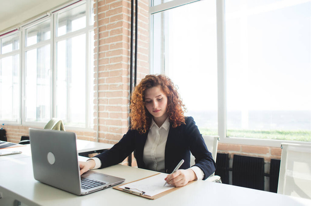 Beautiful young red-haired woman in a business suit works in an office with a laptop - Fotoğraf, Görsel