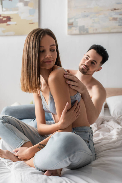 sexy man seducing pretty young woman sitting on bed in jeans and bra - Zdjęcie, obraz