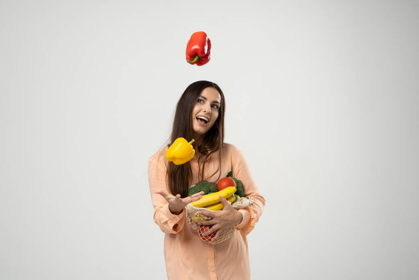 Eco friendly smiling woman in orange dress throw up a red and yellow pepper in a air by one hand and holding reusable cotton eco bags full of groceries in other hand - Foto, imagen