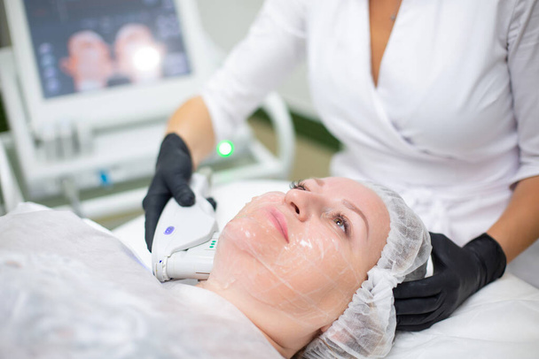 A woman in a cosmetology office receives a SMAS lifting procedure. Non-surgical ultrasound facelift, rejuvenation, moisturizing, getting rid of wrinkles, skin tone and elasticity - 写真・画像