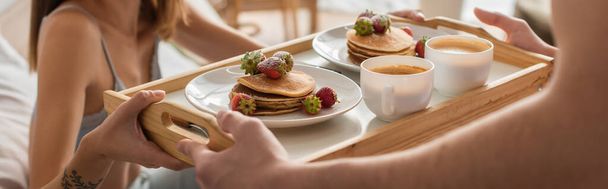 cropped view of blurred man holding tray with coffee, pancakes and fresh strawberries near woman in bedroom, banner - Photo, image