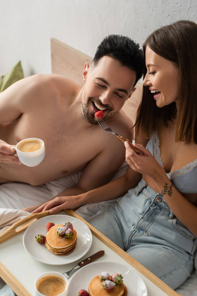 cheerful woman in jeans and bra feeding boyfriend with strawberries during breakfast in bedroom - Photo, image