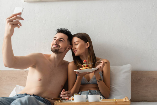 shirtless man taking selfie with pleased girlfriend eating delicious breakfast in bed - Foto, afbeelding