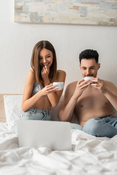 happy and sexy couple drinking morning coffee while watching film on laptop in bedroom - Foto, afbeelding