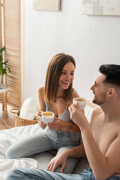 sexy woman and shirtless man smiling at each other while drinking coffee in bedroom - Photo, image