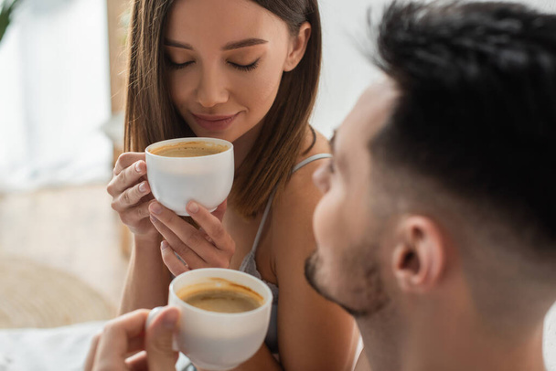sexy woman with closed eyes enjoying morning coffee near blurred boyfriend in bedroom - Foto, afbeelding