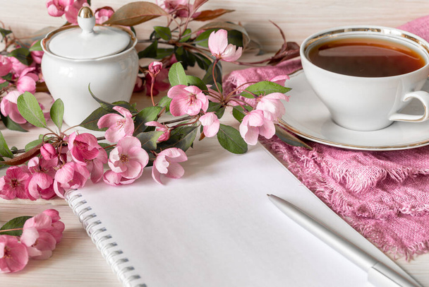 Ein Zweig eines blühenden Apfelbaums, ein leeres Notizbuch, ein Stift und eine Tasse Tee auf einem Holztisch. Den Tag planen oder für den Morgentee einkaufen. Kopierraum - Foto, Bild