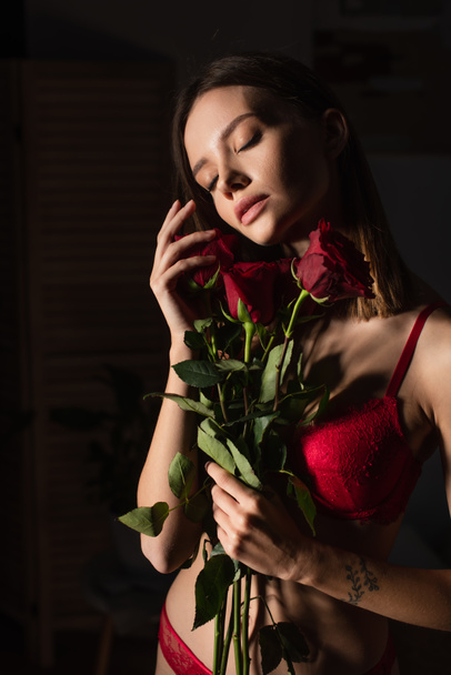 passionate woman in red underwear standing with closed eyes and roses on dark background - 写真・画像