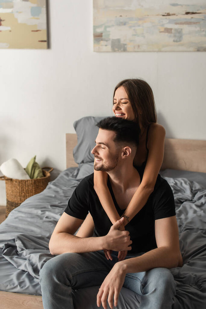 cheerful woman smiling with closed eyes while hugging man sitting on bed and holding her hands - Photo, Image