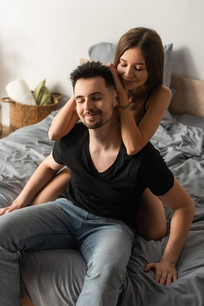 happy woman embracing head of smiling man sitting on bed with closed eyes - Foto, afbeelding