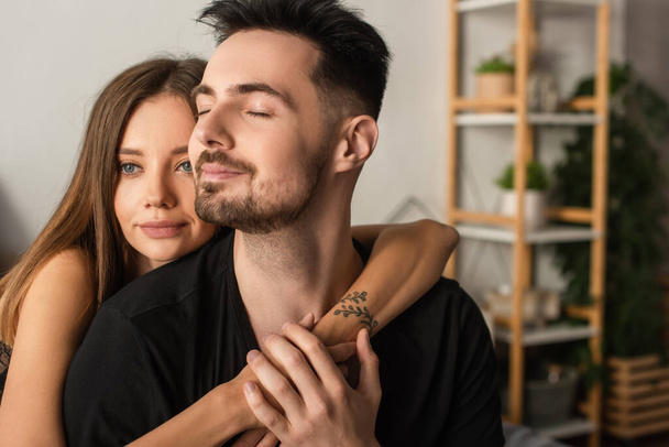 happy tattooed woman hugging boyfriend in black t-shirt sitting with closed eyes in bedroom - Fotoğraf, Görsel