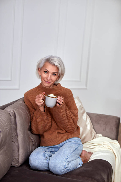 Mujer alegre sentada con taza de té en un cómodo sofá - Foto, imagen