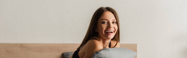 happy seductive woman looking at camera while hugging grey pillow in bedroom, banner - Foto, Bild