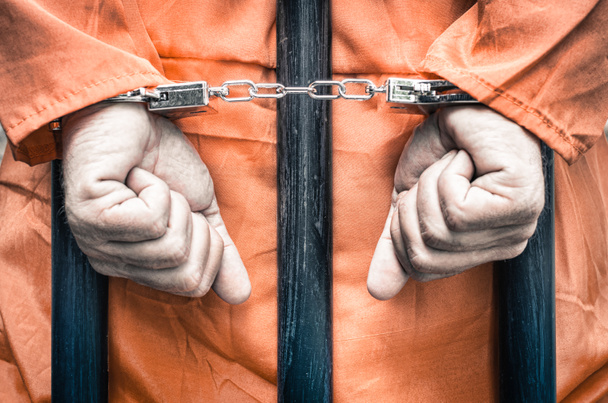 χειροπέδες τα χέρια ενός φυλακισμένου πίσω από τις μπάρες μιας φυλακής με πορτοκαλί ρούχα - τραγανό desaturated βλέμμα δραματική φιλτραρισμένα - Φωτογραφία, εικόνα