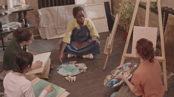 Hochwinkel der inspirierten Afroamerikanerin mit Schürze, auf dem Boden im Kunstatelier sitzend, abstraktes Bild mit Farbe und Pinsel malend, Unterricht und Gespräche mit erwachsenen multiethnischen Studenten - Filmmaterial, Video