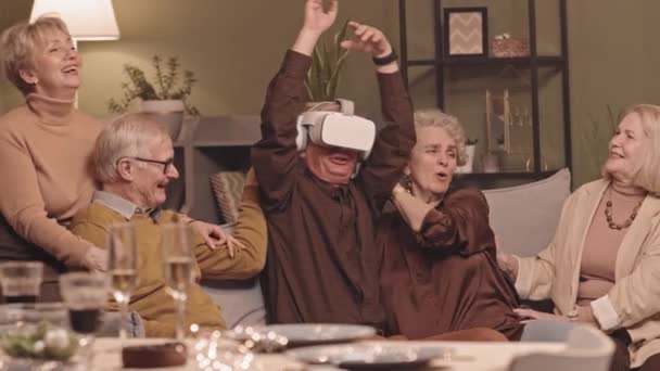 Medium slowmo shot of joyful senior man experiencing virtual reality in headset together with his friends sitting on sofa in cozy living room - Footage, Video