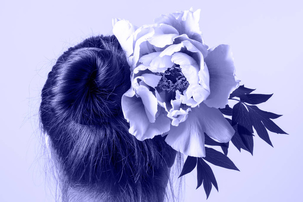 Weiblicher Kopf mit Pfingstrosenblüte im dunklen Haar, Rückansicht. Tonung in der Farbe des Jahres 2022, sehr peri. Trendfarben-Konzept. - Foto, Bild
