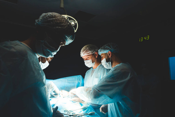 An international professional team of surgeon, assistants and anesthesiologist perform a complex operation on a patient under general anesthesia. Dark atmospheric photography theme in low key. - Фото, изображение