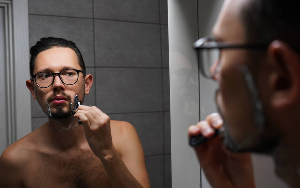 junger gutaussehender Mann mit Brille rasiert sich vor der Arbeit im Badezimmer. junger erfolgreicher Geschäftsmann geht morgens zur Arbeit - Foto, Bild