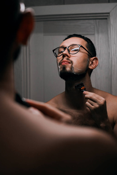 jeune homme beau avec des lunettes se rase dans la salle de bain avant le travail. jeune homme d'affaires réussi va travailler dans la matinée - Photo, image