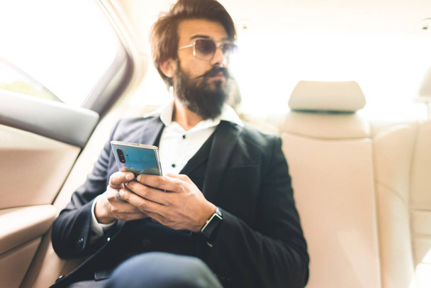 Stylish handsome Hindu boss with glasses and a beard works with the phone in the back seat of the car. Safe and comfortable business travel - Fotoğraf, Görsel