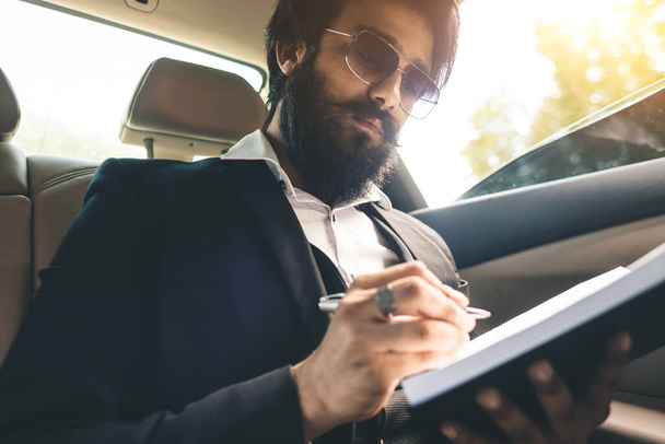 A stylish and successful Indian with glasses and a beard works and plans his schedule in the back seat of the car. Safe and comfortable business travel - 写真・画像