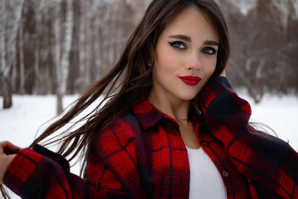 Urocza kobieta z czerwonymi ustami i w szachownicy dotykająca włosów stojąc w śnieżnym lesie zimowym i patrząc w kamerę - Zdjęcie, obraz