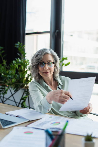 Mature businesswoman holding documents near blurred laptop and notebook in office  - Фото, зображення