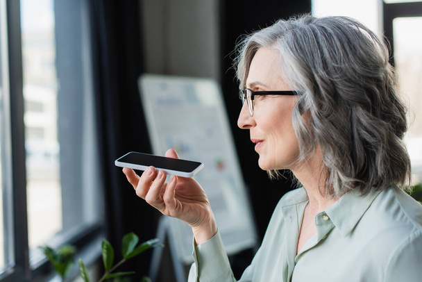 Zijaanzicht van volwassen zakenvrouw in bril opnemen spraakbericht op smartphone in het kantoor  - Foto, afbeelding