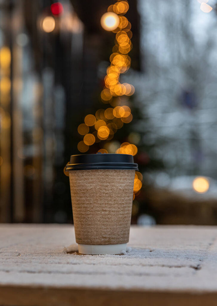 Takeaway disposable cup for hot drink on wooden table. Street walk cafe. Coffee, tea or mulled wine to go. Empty space for text, mockup. Blurred background with garland. - Foto, Imagem