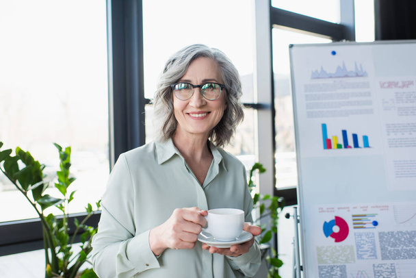 Positive businesswoman looking at camera while holding cup near flip chart in office  - Фото, изображение