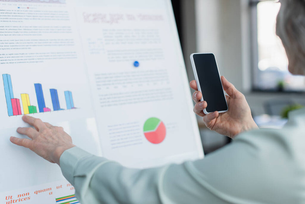 Cropped view of mature businesswoman taking photo on cellphone near flip chart with graphs  - Fotoğraf, Görsel