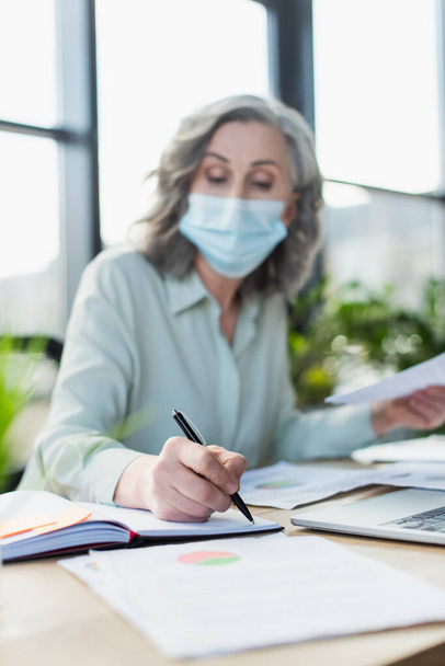 Blurred businesswoman in medical mask writing on notebook near laptop in office  - Fotoğraf, Görsel