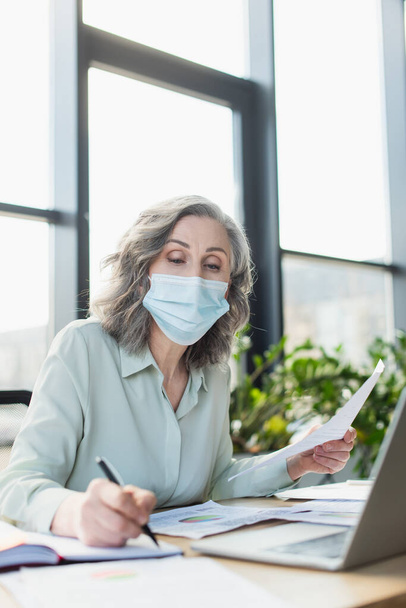Grey haired businesswoman in medical mask holding paper and writing on notebook near laptop in office  - Фото, изображение