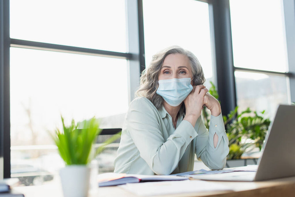 Mature businesswoman in medical mask looking at camera near laptop in office  - Foto, imagen
