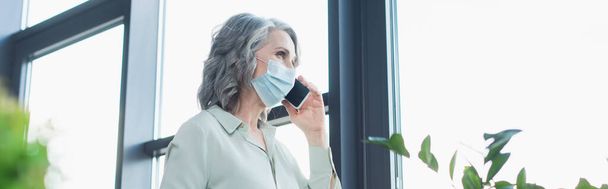 Mujer de negocios de pelo gris en máscara médica hablando en el teléfono inteligente cerca de la ventana en la oficina, pancarta  - Foto, Imagen